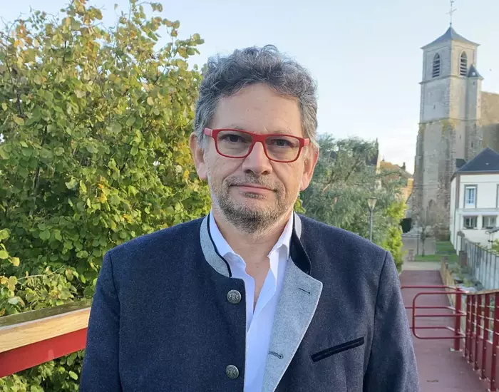 L'image montre un homme de face avec des lunettes noires et un costume rayé gris